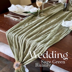 Op de afbeelding: Saliegroene bruidsloper over een donkerhouten tafel. De getextureerde loper is geplooid en heeft een bloemaccent. De tafel is gedekt met glaswerk, bestek en gevouwen servetten. Tekst op de loper luidt "Wedding Sage Green Runner."