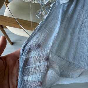 Puede incluir: Un mantel de mesa de tela transparente azul claro con tejido texturizado. La tela est&aacute; drapeada sobre una mesa y sostenida por una mano.