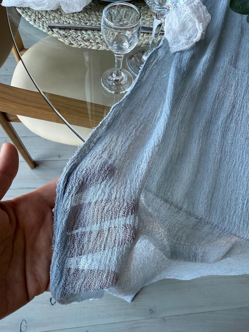 Puede incluir: Un mantel de mesa de tela transparente azul claro con tejido texturizado. La tela est&aacute; drapeada sobre una mesa y sostenida por una mano.