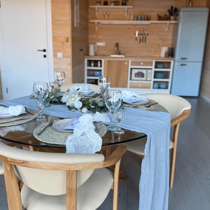 Puede incluir: Una mesa redonda de vidrio con un mantel de mesa azul claro y servilletas blancas. La mesa est&aacute; puesta con cuatro cubiertos, cada uno con un plato blanco, un mantel individual tejido y un vaso. Hay flores y vegetaci&oacute;n en el centro de la mesa. La mesa est&aacute; en un comedor con paredes de madera y un refrigerador azul claro en el fondo.