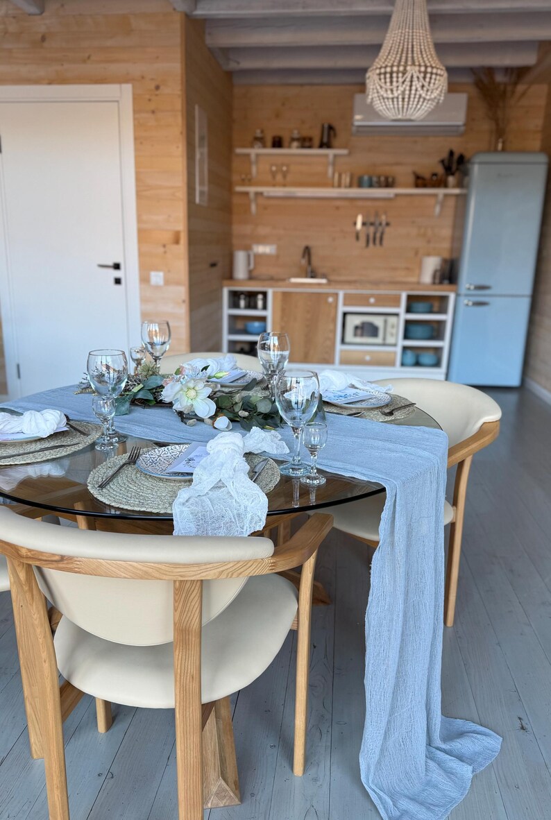 Puede incluir: Una mesa redonda de vidrio con un mantel de mesa azul claro y servilletas blancas. La mesa est&aacute; puesta con cuatro cubiertos, cada uno con un plato blanco, un mantel individual tejido y un vaso. Hay flores y vegetaci&oacute;n en el centro de la mesa. La mesa est&aacute; en un comedor con paredes de madera y un refrigerador azul claro en el fondo.