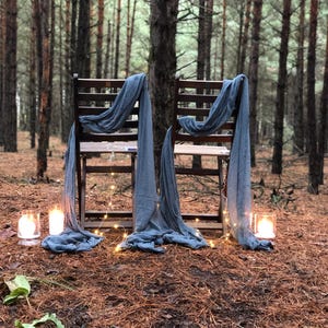 Puede incluir: Dos sillas de madera con tela azul drapeada sobre ellas, instaladas en un bosque de pinos con luces de cadena y velas.