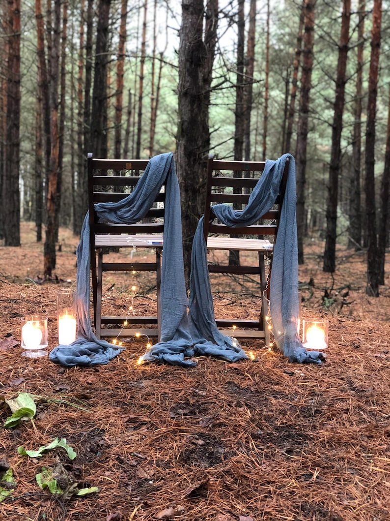 Puede incluir: Dos sillas de madera con tela azul drapeada sobre ellas, instaladas en un bosque de pinos con luces de cadena y velas.