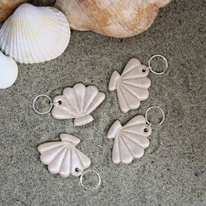 Peut inclure: Quatre porte-clés en forme de coquillage beige avec des anneaux de clés argentés sur un fond de sable. Les porte-clés sont disposés en demi-cercle.