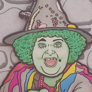 May include: A cartoon illustration of a green-skinned witch wearing a grey pointed hat with pink and yellow accents. She has green curly hair and is wearing a colourful striped jacket. She is pointing at the viewer with her right hand and has a big smile on her face.