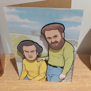 May include: Greeting card featuring a cartoon illustration of two men. The men are wearing yellow shirts and blue jeans. The background shows a blue sky with clouds. The card is made of cardboard and is displayed on a wooden shelf.