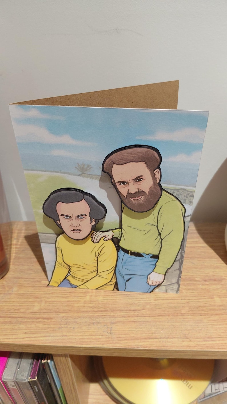 May include: Greeting card featuring a cartoon illustration of two men. The men are wearing yellow shirts and blue jeans. The background shows a blue sky with clouds. The card is made of cardboard and is displayed on a wooden shelf.