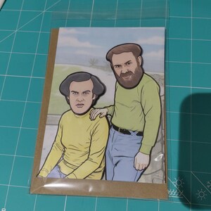 May include: A greeting card featuring a cartoon illustration of two men in yellow shirts and blue jeans. One man has a beard and is placing his hand on the other man's shoulder. The background shows a landscape with a blue sky.