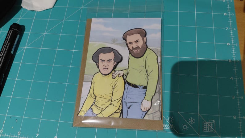 May include: A greeting card featuring a cartoon illustration of two men in yellow shirts and blue jeans. One man has a beard and is placing his hand on the other man's shoulder. The background shows a landscape with a blue sky.