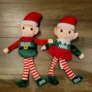 Può includere: Due bambole di elfo di peluche con gambe e cappelli a righe rosse e bianche. L'elfo a sinistra indossa un vestito verde con il nome "Derek" ricamato. L'elfo a destra indossa un vestito rosso con il nome "Anna" ricamato. Entrambi gli elfi hanno il 2023 ricamato sulle loro scarpe.