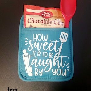 Może przedstawiać: Turkusowa rękawica do piekarnika z napisem "HOW sweet it is to be taught by you" w kolorze białym. Wewnątrz rękawicy znajduje się czerwona łyżka i opakowanie mieszanki do ciasteczek z kawałkami czekolady Betty Crocker.