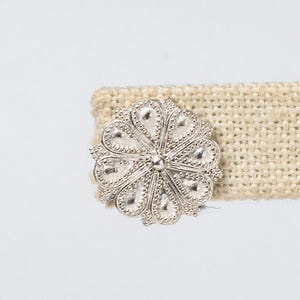 May include: A silver flower-shaped button with a textured design, attached to a beige fabric strap.