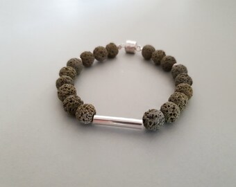 May include: A bracelet featuring olive green lava rock beads and a silver-colored cylindrical accent. The bracelet is strung on a cord and has a magnetic clasp. The beads have a porous texture.