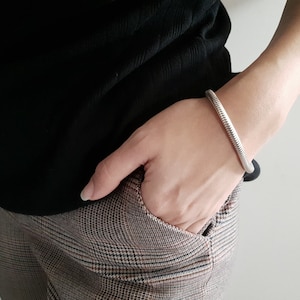 Puede incluir: Un brazalete plateado texturizado. El brazalete tiene un diseño acanalado y se usa en la muñeca. La persona lleva una camiseta negra y pantalones a cuadros.