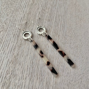 May include: Dangle earrings featuring a silver-coloured circular top and a long, rectangular pendant. The pendant has a tortoiseshell pattern in cream, black, and brown. The earrings are presented on a light-coloured wooden surface.