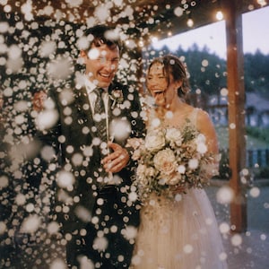 May include: A person in a dark suit with a patterned tie and a person in a light-coloured sleeveless gown holding a bouquet of white and pink flowers. They are laughing while surrounded by falling white confetti under string lights at dusk.