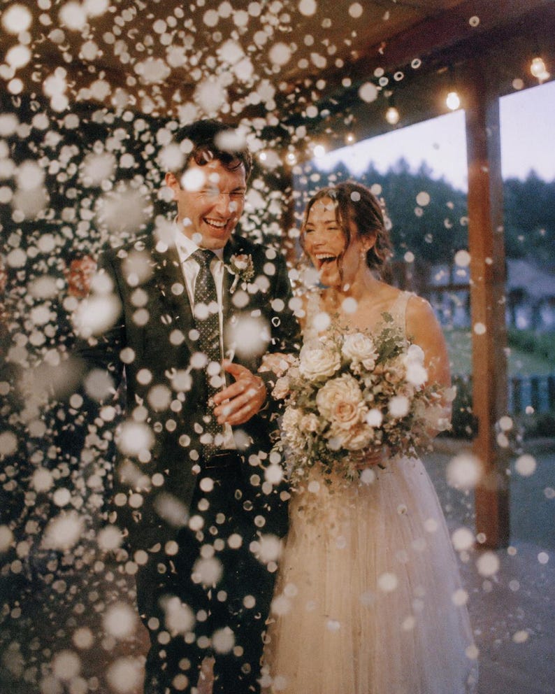 May include: A person in a dark suit with a patterned tie and a person in a light-coloured sleeveless gown holding a bouquet of white and pink flowers. They are laughing while surrounded by falling white confetti under string lights at dusk.