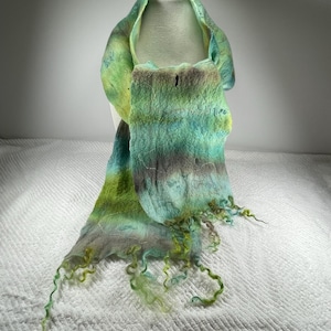 Può includere: Sciarpa in feltro fatta a mano nei toni del verde, blu e grigio. La sciarpa ha un aspetto strutturato con un bordo sfrangiato. I colori si fondono in strisce orizzontali, creando un accessorio unico e artistico.