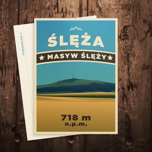 Peut inclure: Une carte postale de voyage avec un paysage de montagne. Le texte "ŚLĘŻA" et "MASYW ŚLĘŻY" en blanc se détachent sur un ciel bleu. La carte postale affiche également la mesure "718 m n.p.m." et est posée sur un fond en bois.