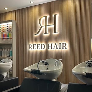 以下が含まれることがあります： 木製のスラット壁のある美容院の内観。サロンのロゴ「RH REED HAIR」は金色に照らされています。白い洗面台と黒い椅子が見え、プロのヘアケア環境を示唆しています。