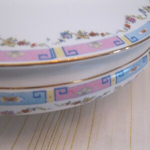 May include: A white ceramic cake stand with a pink, blue, and yellow geometric pattern around the edge. The stand has a gold trim.