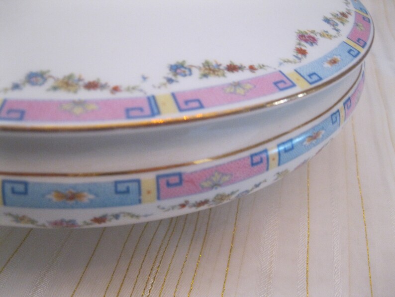 May include: A white ceramic cake stand with a pink, blue, and yellow geometric pattern around the edge. The stand has a gold trim.