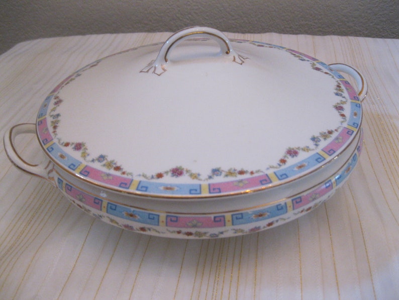 May include: White porcelain serving dish with a lid. The dish has a floral pattern with pink, blue, and yellow accents. The lid has a gold trim and a single handle.
