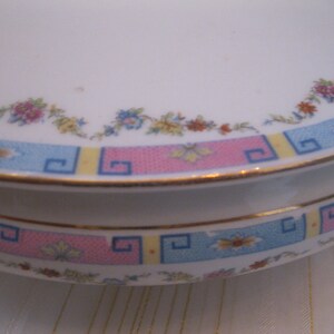 May include: A white ceramic dish with a pink, blue, and yellow geometric pattern around the rim. The dish has a floral design with pink, yellow, and blue flowers.