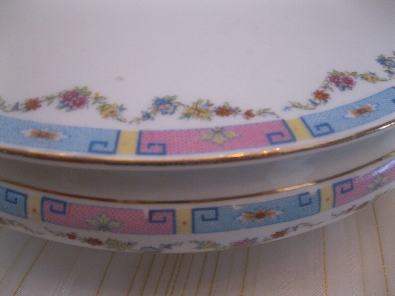 May include: A white ceramic dish with a pink, blue, and yellow geometric pattern around the rim. The dish has a floral design with pink, yellow, and blue flowers.