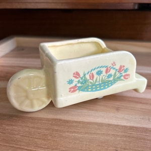 May include: A small, yellow ceramic planter shaped like a wheelbarrow with a floral design. The planter has a blue and white floral pattern with pink tulips and green leaves.