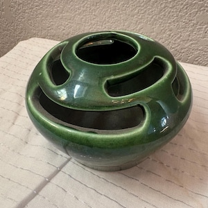 May include: A round, dark green ceramic bowl with a glossy finish and a unique top design featuring three large, curved cutouts. The bowl has a wide, open center and a smooth, rounded exterior surface that tapers toward the base, creating a distinct shape.