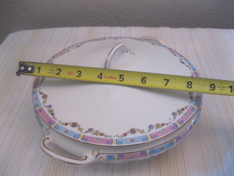 May include: White ceramic serving dish with a floral design in pink, blue, and gold. The dish has two handles and a lid with a handle. The dish measures 9 inches in diameter.