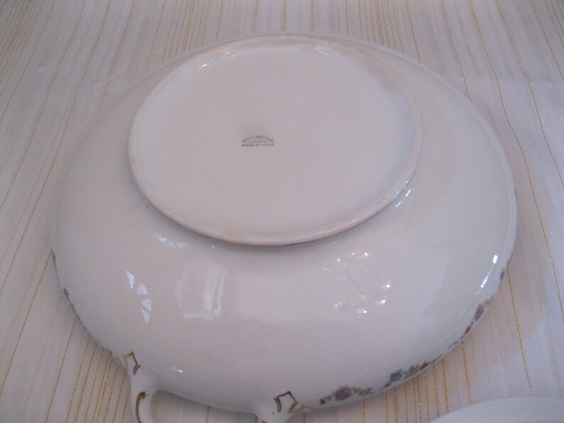 May include: White ceramic serving bowl with a floral pattern and gold trim. The bowl has two handles and a raised center section. The bottom of the bowl has the text "Made in U.S.A."