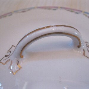 May include: A white porcelain lid with a gold-trimmed handle. The handle is shaped like an arch and has a decorative design on each end.