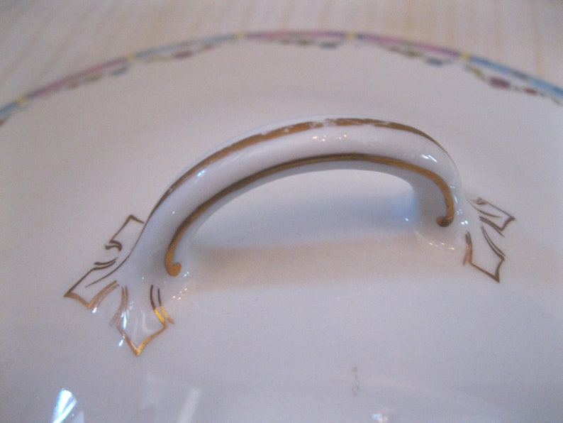 May include: A white porcelain lid with a gold-trimmed handle. The handle is shaped like an arch and has a decorative design on each end.