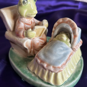 May include: A ceramic figurine depicting a frog reading to a baby frog in a bassinet. The frog is wearing a peach-colored outfit and seated in a chair. The bassinet is light blue with a peach trim, resting on a green base.
