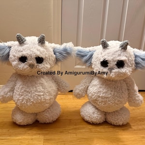 May include: Two white plush creatures with fuzzy bodies, large black eyes, small grey horns, and light blue faux fur ears. The text Created By AmigurumiByAmy is centered between the two figures. Each creature has a round body and limbs.
