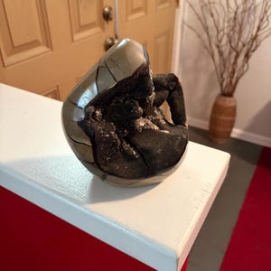 May include: A polished, spherical stone geode featuring a smooth, metallic-toned outer shell with visible cracks and a deep, dark, textured interior cavity filled with small, crystalline formations and granular, black mineral deposits.