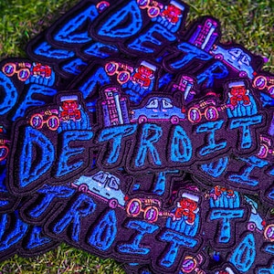 Könnte beinhalten: Eine Ansammlung von gestickten Aufnähern mit dem Wort "DETROIT" in blauen, stilisierten Buchstaben. Die Aufnäher zeigen Illustrationen von Autos, Gebäuden und Figuren. Die Aufnäher befinden sich auf einem schwarzen Hintergrund mit blauem Rand.