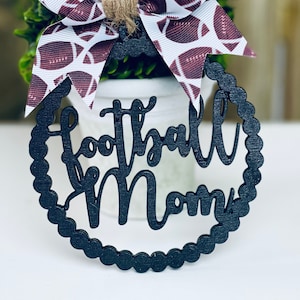 Puede incluir: Adorno de madera negro con las palabras "Football Mom" en un c&iacute;rculo blanco con una cinta marr&oacute;n y blanca con balones de f&uacute;tbol impresos.