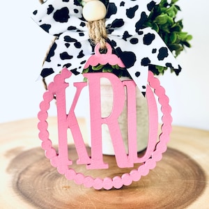 May include: Pink wooden monogram ornament with the letters "KRD" in a circle with a white cow print bow and wooden beads.