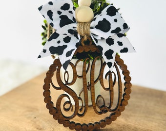 May include: A wooden monogram ornament with a scalloped edge, featuring a three-letter monogram. It is adorned with a black and white cow print bow and a wooden bead. The ornament hangs from a jute twine.