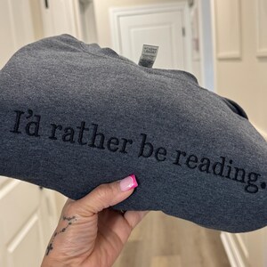 K&ouml;nnte beinhalten: Dunkelgraues Sweatshirt mit dem gestickten Text "I'd rather be reading." Das Sweatshirt ist gefaltet. Der Text ist in einer einfachen Schriftart gehalten. Der Hintergrund ist neutral. Die Abbildung zeigt eine Person, die das Sweatshirt h&auml;lt.