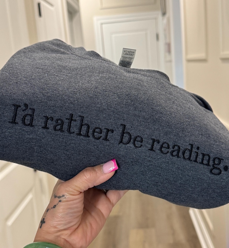 K&ouml;nnte beinhalten: Dunkelgraues Sweatshirt mit dem gestickten Text "I'd rather be reading." Das Sweatshirt ist gefaltet. Der Text ist in einer einfachen Schriftart gehalten. Der Hintergrund ist neutral. Die Abbildung zeigt eine Person, die das Sweatshirt h&auml;lt.