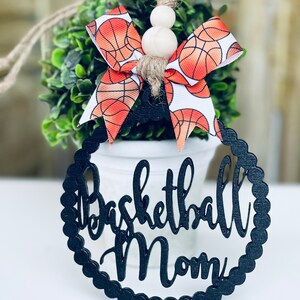 Puede incluir: Adorno de madera negro con las palabras "Basketball Mom" en una fuente decorativa. El adorno est&aacute; decorado con un lazo de baloncesto blanco y naranja.