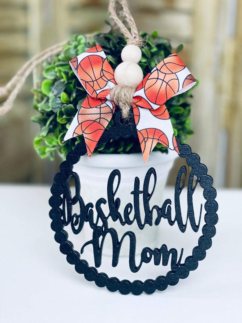 Puede incluir: Adorno de madera negro con las palabras "Basketball Mom" en una fuente decorativa. El adorno est&aacute; decorado con un lazo de baloncesto blanco y naranja.