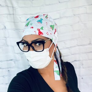May include: A white scrub hat with a Christmas gnome pattern. The hat has a button and ties on the side. The person wearing the hat is also wearing black framed glasses and a white face mask.