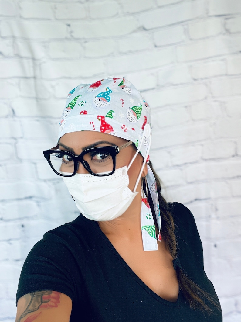 May include: A white scrub hat with a Christmas gnome pattern. The hat has a button and ties on the side. The person wearing the hat is also wearing black framed glasses and a white face mask.