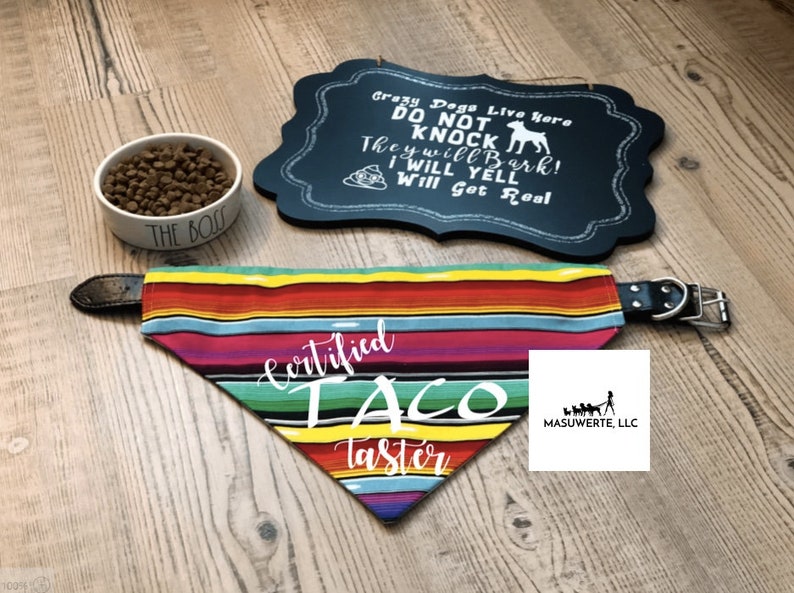 May include: A colorful dog bandana with the text "Certified Taco Taster" in white. The bandana is a traditional Mexican serape pattern with red, yellow, green, blue, and pink stripes. The bandana is attached to a black leather dog collar.