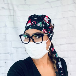 May include: A woman wearing a black scrub cap with a Christmas gnome pattern, black framed glasses, a white face mask, and a black shirt. Her hair is braided and tied with a matching Christmas gnome patterned fabric.